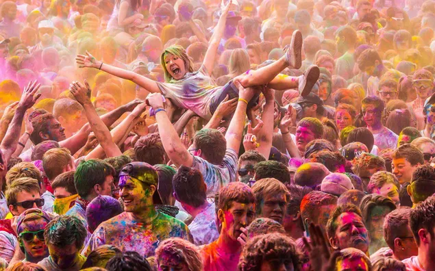 Muat turun Orang ramai yang dicat berwarna-warni dan seorang wanita dilemparkan ke udara semasa perayaan Holi, yang meraikan ketibaan musim bunga