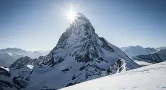 a majestic, snow-covered mountain peak rises sharply under a clear blue sky, illuminated by the bright sun and surrounded by breathtaking alpine scene download