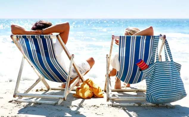 Ein Mann und eine Frau liegen auf einer Chaiselongue im Sand und beobachten die Sonnenstrahlen, die auf das Meer treffen 2K Hintergrundbild