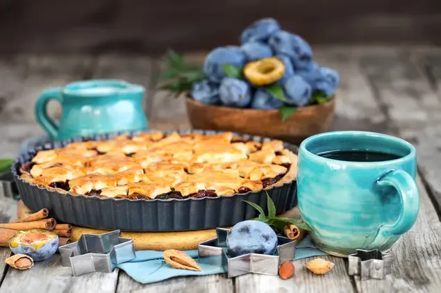 Pai plum utuh dengan batang kayu manis dan buah prem dengan secangkir kopi hitam di sampingnya