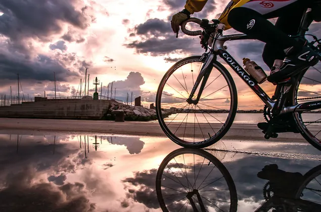 Atleet wat fiets ry en die weerkaatsing van die fiets in die water met wolke aflaai