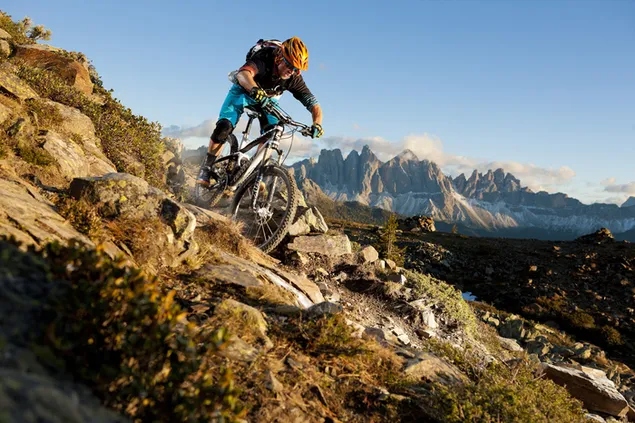 Muat turun Atlet dengan beg galas dalam topi keledar menunggang basikal gunung di atas jalan tanah yang kasar di puncak