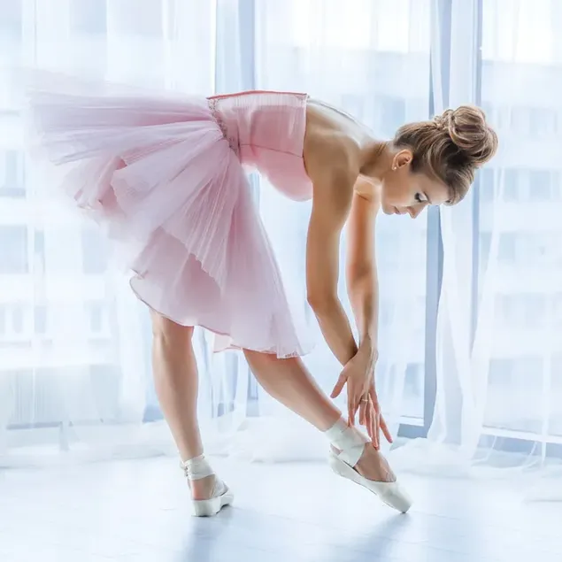 Beautiful ballerina girl in white ballerina shoes in pink dress dancing near the white curtains in front of the window