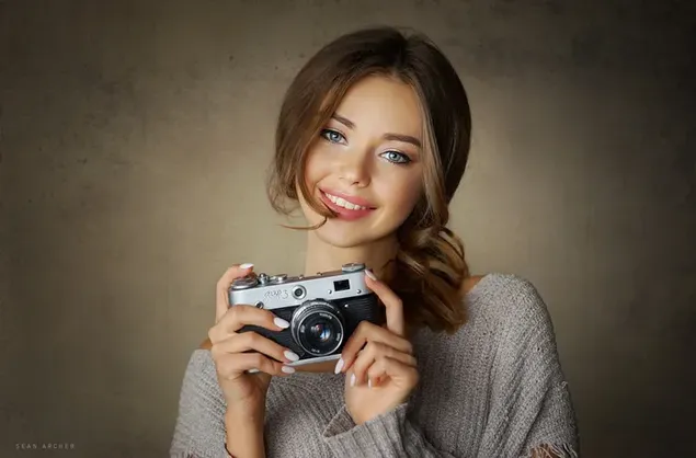 Beautiful female model with chestnut hair, gray sweater and blue eyes holding camera in front of brown background