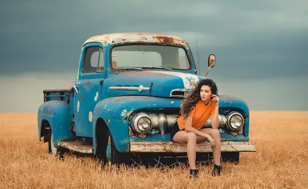 Model wanita cantik dengan rambut keriting berpose dengan truk pickup biru berkarat yang diparkir di luar ruangan