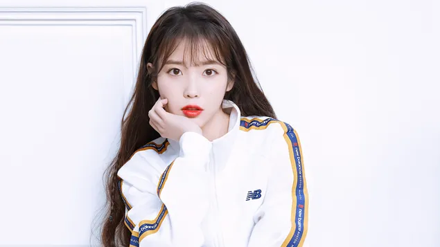 Beautiful singer IU poses in front of a white background with her hands on her chin, long brown hair in a white dress.