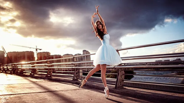 Mooie vrouw in witte jurk die ballet doet in donkere bewolkte stad tussen zee, brug en gebouwen.