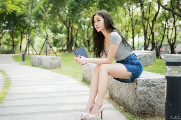 Chica asiática de cabello negro sentada en un banco de roca en el parque 4K fondo de pantalla