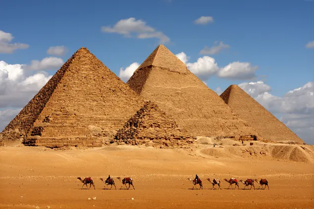 Camels and people travel on desert sand in front of cloudy sky and Egyptian pyramids