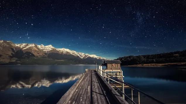 Camino de madera y cabaña en el lago con estrellas reflejadas y montañas nevadas por la noche