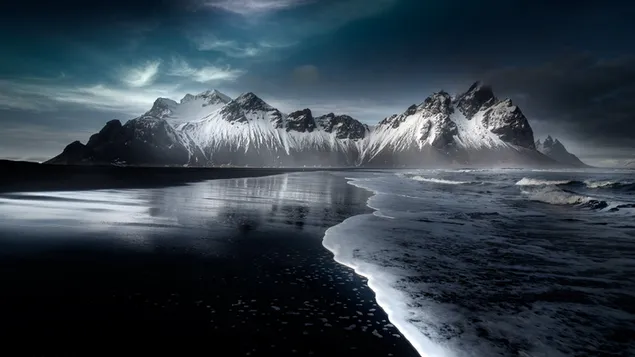 Kombination aus schneebedeckten Bergen und Meereswellen auf schwarzem Sand 4K Hintergrundbild
