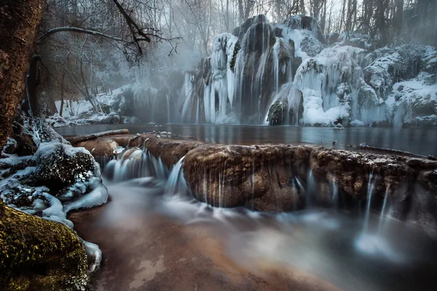 De natuurlijke stroom van de waterval die tussen de bomen in het bos stroomt, in de sneeuw en mist