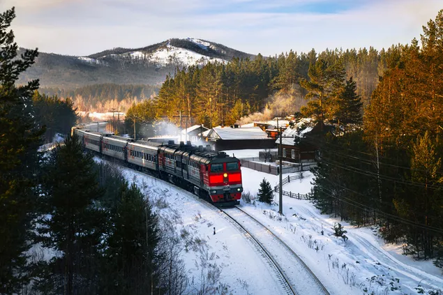 Departure of train from red train station moving on railway between snowy mountains and forests
