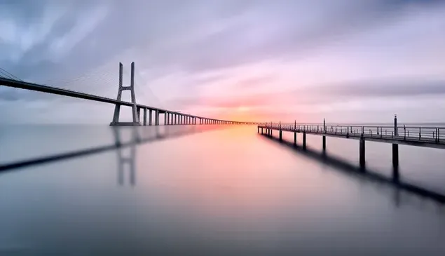 Der Blick auf die Sonne, die zwischen den Wolken im Freien aufgeht und die Brücke über dem Meer beleuchtet herunterladen