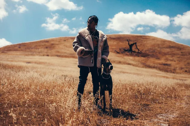 El rapero y productor estadounidense Travis scott posando con su perro en un campo de tierra con una vista de nubes