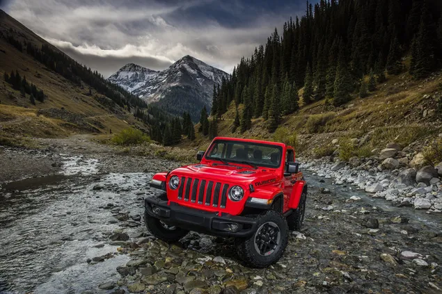 Fahrzeug der Marke Jeep mit seiner faszinierenden roten Farbe auf einer unbefestigten Straße zwischen Wäldern und schneebedeckten Bergen herunterladen