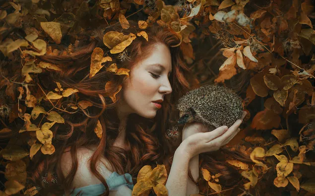 Female model with long red hair among autumn leaves holding a cute hedgehog