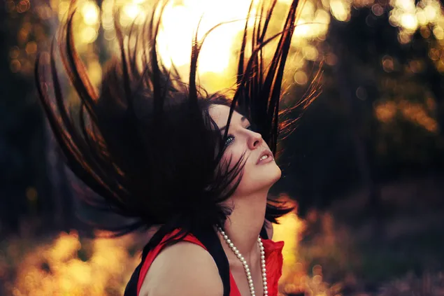 Gaze of beautiful woman with blue eyes and pearl necklace, in red dress, tossing her hair in front of blurry forest backdrop