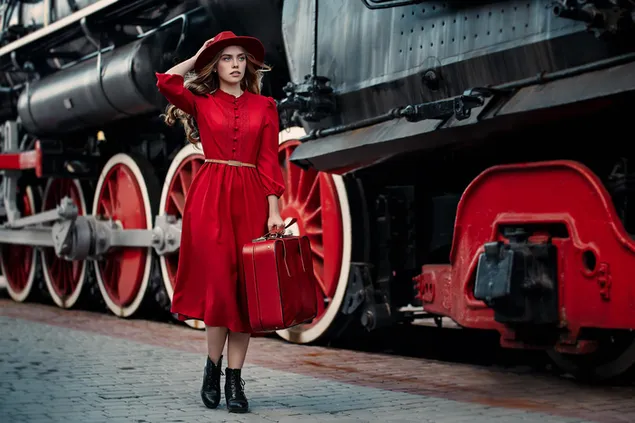 Girl in red dress holding a suitcase in hands beside vintage train