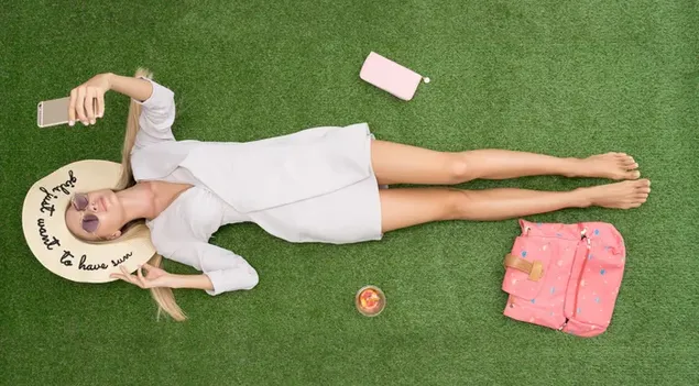 Tapeta Dívka, která nosí bílé šaty a letní klobouk drží telefon a leží na trávě 4K