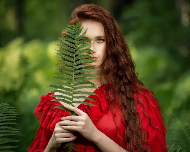 Hermosa modelo femenina con cabello castaño largo, ojos verdes, vestido rojo sosteniendo hojas de pino frente a un fondo verde borroso