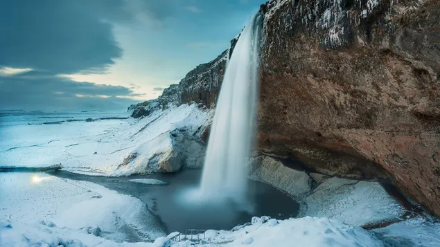 Het uitzicht op de waterval die in de winter van de rotsen in de natuur onder de sneeuw stroomt