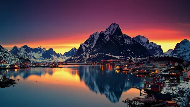 Houses and mountains reflect in the water as the yellow red lights of the sunset rise from under the snowy mountains 2K wallpaper