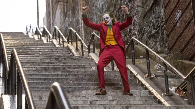Joker dengan riasan uniknya dalam jaket merah dan rompi kuning di tangga di samping dinding yang dicat grafiti 4K wallpaper