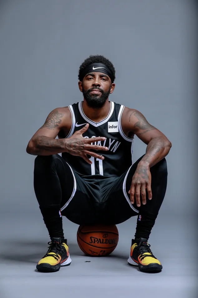 Kyrie irving sitting on a basketball in a brooklyn nets jersey