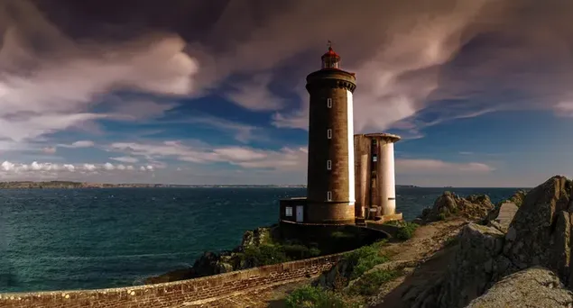Leuchtturm in Frankreich vor Steinen am Ufer des Meeres bei dunklem bewölktem Wetter herunterladen