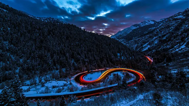Long exposure photo of car lights on asphalt road between snowy mountains and forest