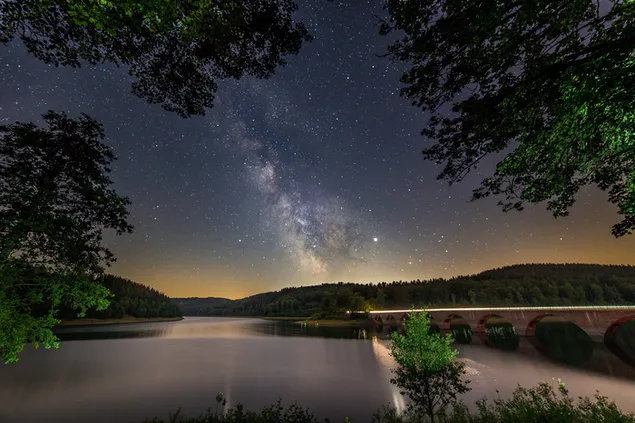 Maravillosa noche estrellada brillante en el cielo vista a través del largo puente y hojas de árboles sobre el lago
