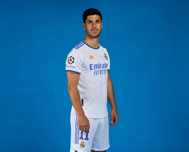 Marco Asensio posa de pie frente a un fondo azul con la camiseta del Real Madrid