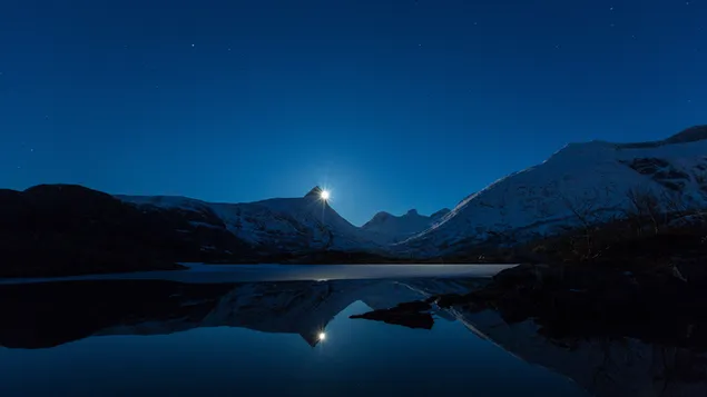 Luz de luna detrás de montañas nevadas y siluetas de montaña reflejadas en el agua