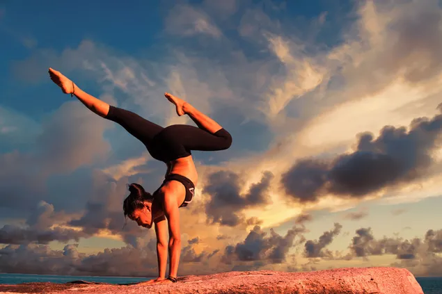 Mujer en ropa deportiva haciendo yoga en roca junto al mar y vista de nubes