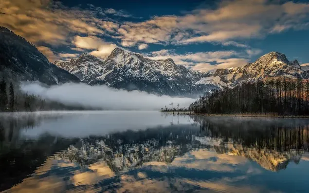 Niebla reunida al pie de montañas y árboles nevados y el reflejo del cielo en el agua del lago