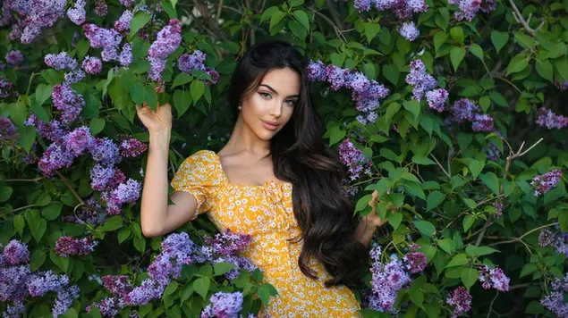 Pose van mooi vrouwelijk model met zwart lang haar in gele jurk tussen paarse bloemen