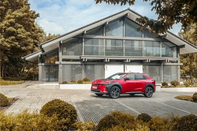 Rote Farbe Toyota bz4x parkte auf Steinpflaster vor zweistöckigem Haus zwischen Bäumen 4K Hintergrundbild
