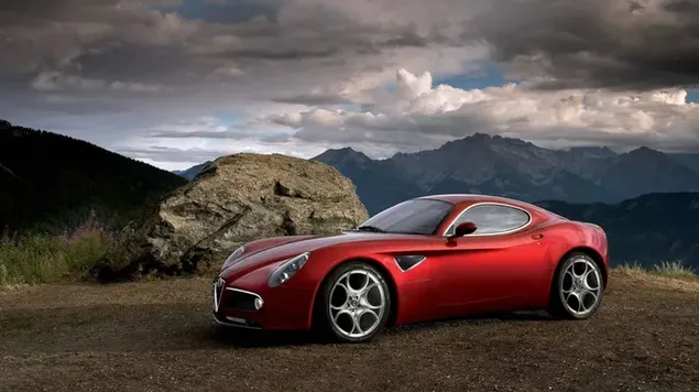 Red colored sports car Alfa Romeo with steel wheels parked on a dirt road among the mountains in cloudy weather