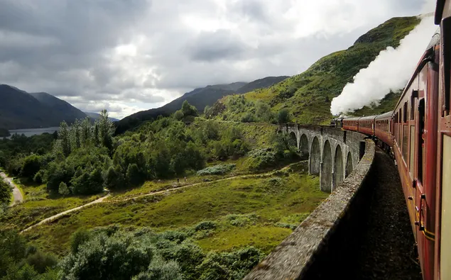 Roter Zug, der sich auf der Brücke in der Naturlandschaft bewegt, die mit Bergen, Felsen und Wolken geschmückt ist 4K Hintergrundbild