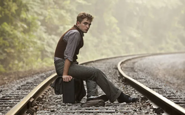 Robert pattinson sitting on his suitcase on the train tracks in the forest