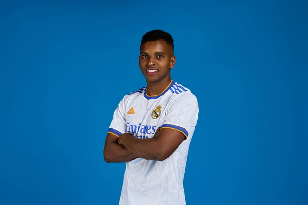 Rodrygo poseert met gevouwen handen voor een blauwe achtergrond, gekleed in de trui van Rebellion La Liga sterk team Real Madrid