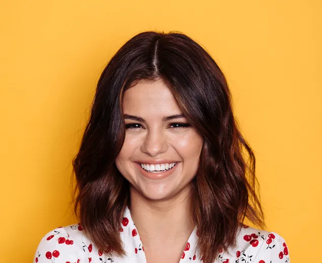 Selena Gomez lächelt, in einem weißen Kleid mit Kirschmuster und gelbem Hintergrund