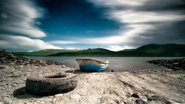 Small boat and old car tire on the river next to distant mountains and dark clouds