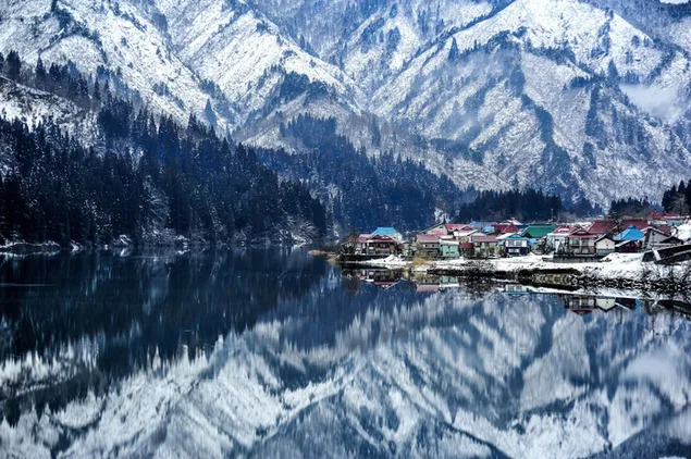 Small town houses reflected in lake water at the foot of snowy mountains and forest trees