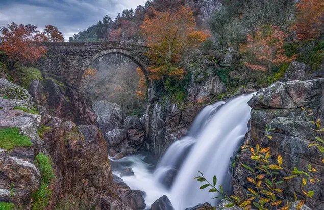 Pont de pierre à côté de la cascade et des feuilles d'arbres séchées en automne par temps nuageux sombre 6K fond d'écran