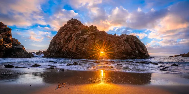 La luz del sol se filtra entre las rocas en medio del mar y el reflejo del cielo nublado en las arenas mojadas de la playa