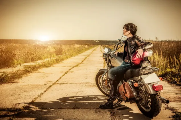 太陽の黄色い光線の石の道の乾いた草の近くのオートバイに座っている編んだ髪の美しい女性