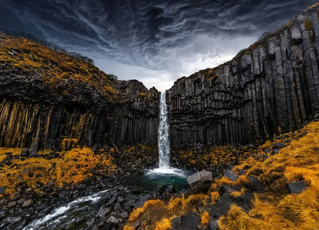 La magnífica cascada que parece fluir a través de las nubes oscuras fluye a través de las rocas hacia la hierba seca y las piedras. HD fondo de pantalla