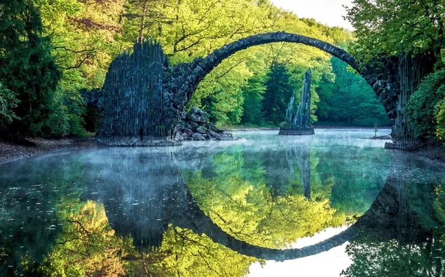 El reflejo del puente y los árboles del bosque en el agua en la foto donde un marco parece dos marcos separados. 2K fondo de pantalla